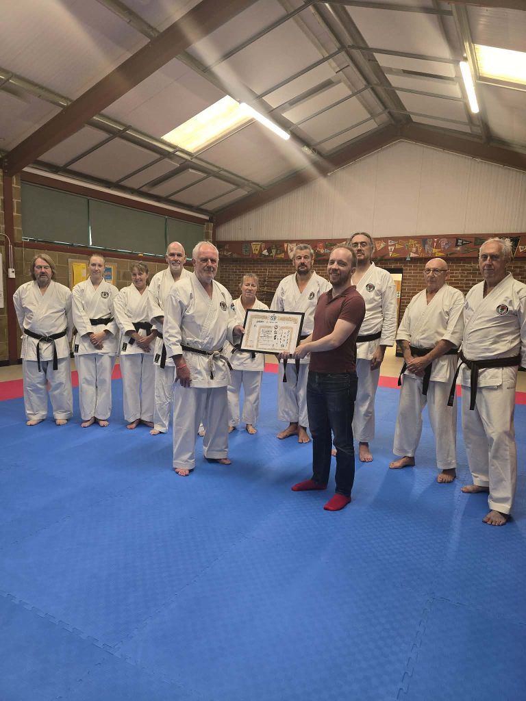 Oren Teichmann being presented with 5th Dan from BWF President Paul Elliott 8th Dan Kyoshi. Both men are stood holding the certificate between them and are surrounded by other black belts members of SEMKA karate.