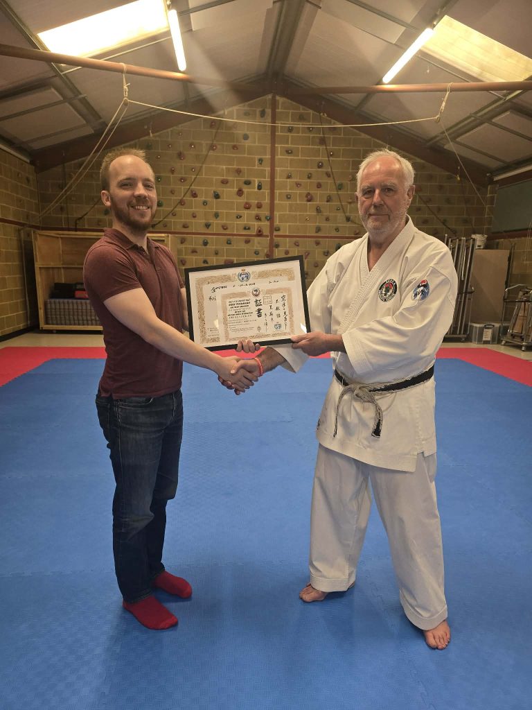 Oren Teichmann receiving 5th Dan from BWF President Paul Elliott 8th Dan Kyoshi. Both men are shaking hands and holding the certificate between them.
